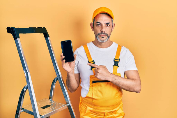 Knappe man van middelbare leeftijd met grijs haar stand-by ladder met smartphone ontspannen met serieuze uitdrukking op het gezicht. eenvoudig en natuurlijk kijkend naar de camera.  - Foto, afbeelding