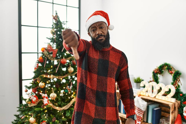 African american man wearing santa claus hat standing by christmas tree looking unhappy and angry showing rejection and negative with thumbs down gesture. bad expression.  - Foto, afbeelding