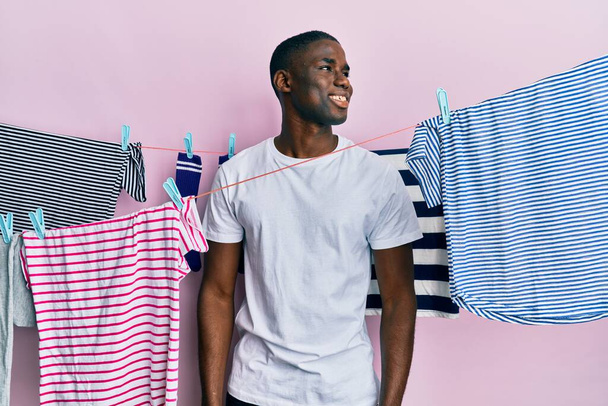 Молодой африканский американец стирает одежду в бельевой веревке глядя в сторону, расслабиться профиль позировать с естественным лицом и уверенной улыбкой.  - Фото, изображение