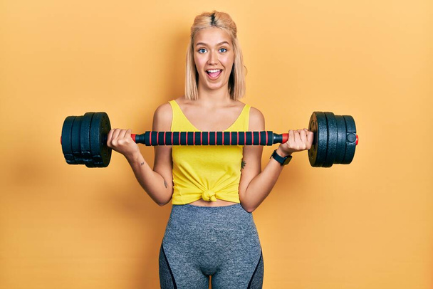 Beautiful blonde woman wearing sportswear using dumbbells celebrating crazy and amazed for success with open eyes screaming excited.  - Photo, Image