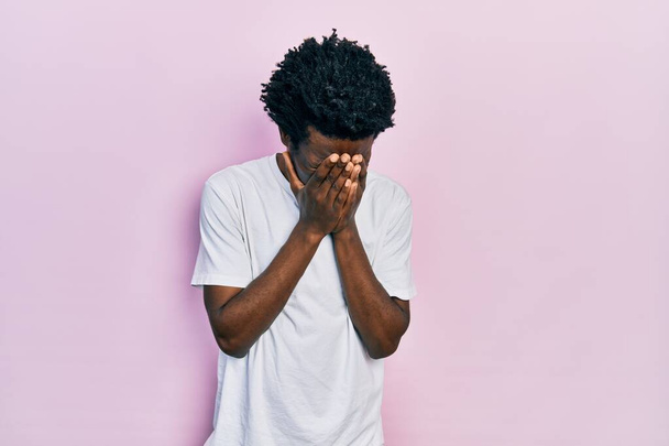 Junger afrikanisch-amerikanischer Mann in lässigem weißem T-Shirt mit traurigem Gesichtsausdruck, der weinend das Gesicht mit den Händen bedeckt. Depressionskonzept.  - Foto, Bild