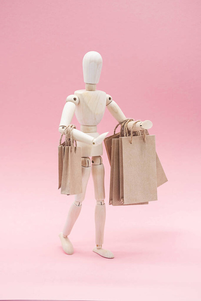 Concetto di vendita o shopping, in stile minimalista. Burattino in legno con borse della spesa artigianali di carta, sullo sfondo rosa. - Foto, immagini