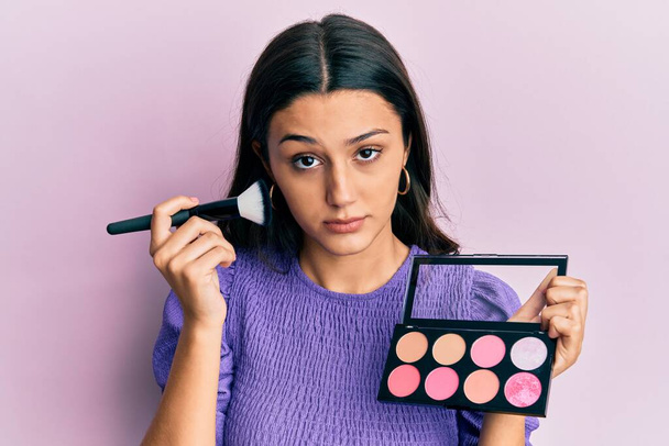Young hispanic woman holding makeup brush and blush skeptic and nervous, frowning upset because of problem. negative person.  - Photo, Image