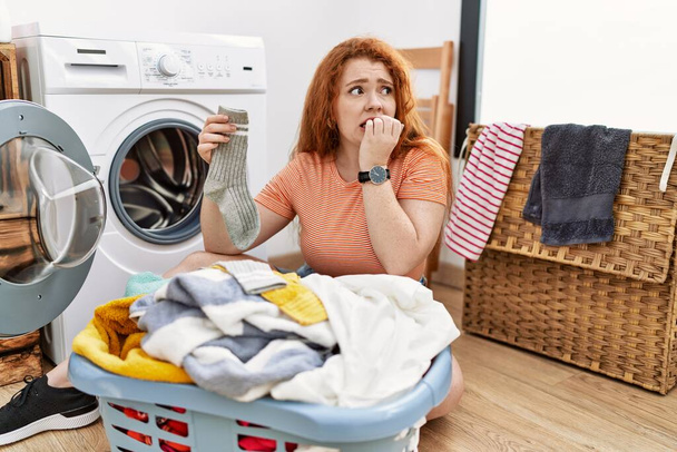 Giovane donna rossa che mette il bucato sporco in lavatrice cercando stressato e nervoso con le mani in bocca mordere le unghie. problema di ansia.  - Foto, immagini