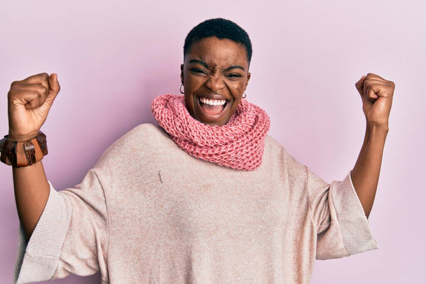 Young african american woman wearing winter scarf screaming proud, celebrating victory and success very excited with raised arms  - Photo, Image
