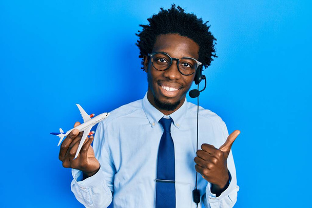 Jonge Afro-Amerikaanse reisagent man houdt vliegtuig glimlachend gelukkig en positief, duim omhoog doet uitstekend en goedkeuring teken  - Foto, afbeelding
