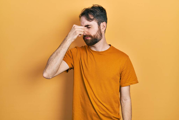 Caucasian man with beard wearing casual yellow t shirt smelling something stinky and disgusting, intolerable smell, holding breath with fingers on nose. bad smell  - Photo, Image