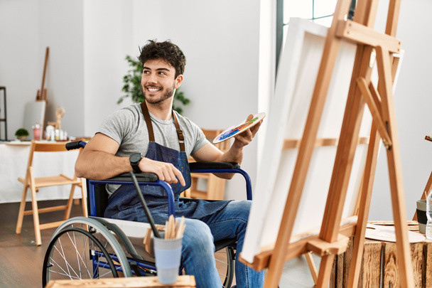 Młody Hiszpan siedzący na wózku inwalidzkim malujący w pracowni sztuki z uśmiechem na twarzy, naturalnym wyrazem twarzy. śmiać się pewnie.  - Zdjęcie, obraz
