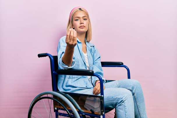 mooie blonde vrouw zitten op rolstoel doen italiaans gebaar met hand en vingers zelfverzekerde uitdrukking  - Foto, afbeelding