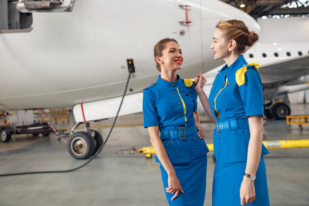 Две красивые стюардессы в стильной ярко-голубой форме улыбаются друг другу, стоя вместе перед самолетом в ангаре в аэропорту - Фото, изображение