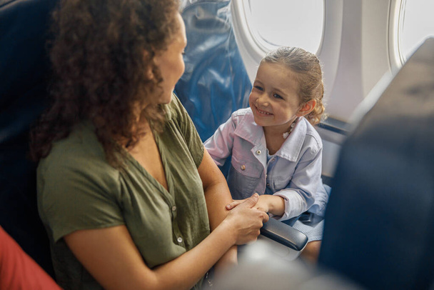 Wysoki kąt widzenia dziewczynki siedzącej w samolocie, uśmiechającej się do matki podczas gdy podróżują razem - Zdjęcie, obraz