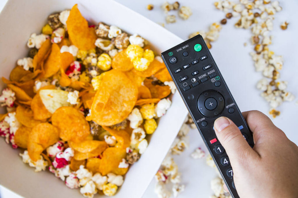 A top view of a hand holding tv remote control against fried popcorn and chips on a bowl - Foto, afbeelding