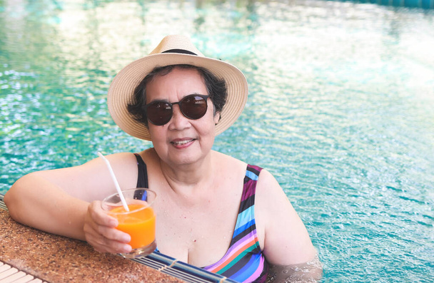 Πορτρέτο της ευτυχισμένης και υγιούς Ασιάτισσας ηλικιωμένης γυναίκας που φοράει πολύχρωμο μαγιό, ψάθινο καπέλο και γυαλιά ηλίου πίνοντας χυμό πορτοκαλιού στην πισίνα, χαμογελώντας και κοιτάζοντας την κάμερα. Ενεργός τρόπος ζωής και ταξιδιωτική έννοια. - Φωτογραφία, εικόνα