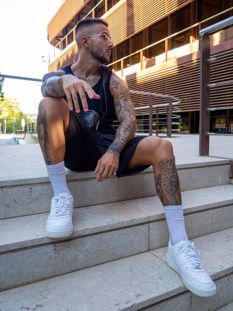 An attractive cool Spanish man with tattoos sitting on the steps in a park on a sunny day - Foto, imagen