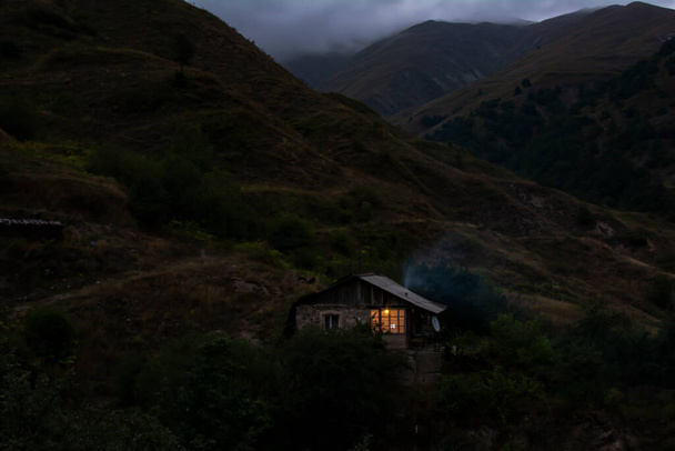 Одинокий домик в горах. Горный пейзаж с небольшим домом. Свет в окне дома. Дым идет из дымохода - Фото, изображение
