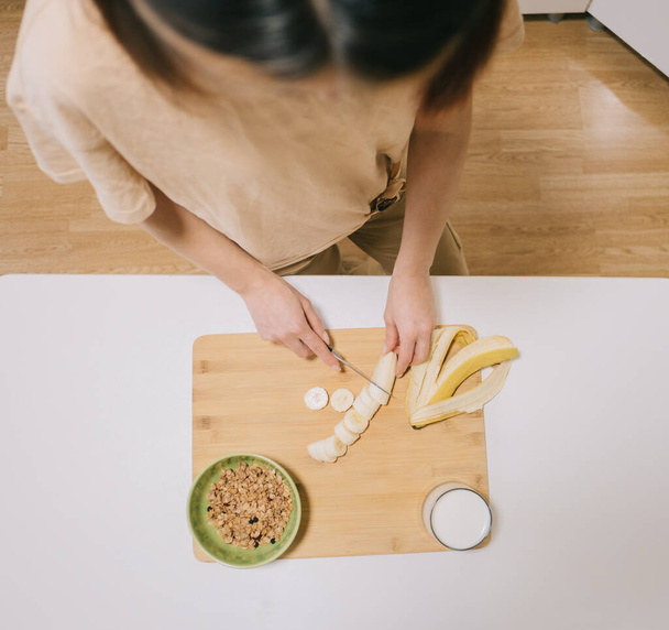 Young woman slices breakfast banana with granola and yogurt. View from above. - 写真・画像