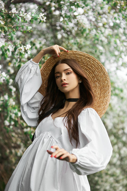 Brown-haired woman in a large straw hat and a white dress poses against the background of blooming white trees. Romantic look, natural beauty, clean facial skin - Photo, image
