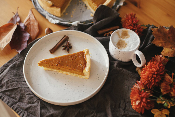 Happy Thanksgiving. Pumpkin pie slice on modern plate and hot coffee on rustic table with linen napkin, autumn flowers and leaves, anise and cinnamon. Homemade pumpkin tart recipe. - Photo, Image