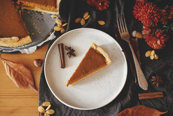 Happy Thanksgiving. Pumpkin pie slice on modern plate on rustic table with linen napkin, autumn flowers and leaves, anise and cinnamon. Homemade pumpkin tart recipe. Top view - Photo, image