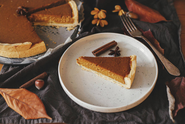 Pumpkin pie slice on modern plate on rustic table with linen napkin, autumn leaves, anise and cinnamon. Happy Thanksgiving. Homemade pumpkin tart recipe - Fotó, kép