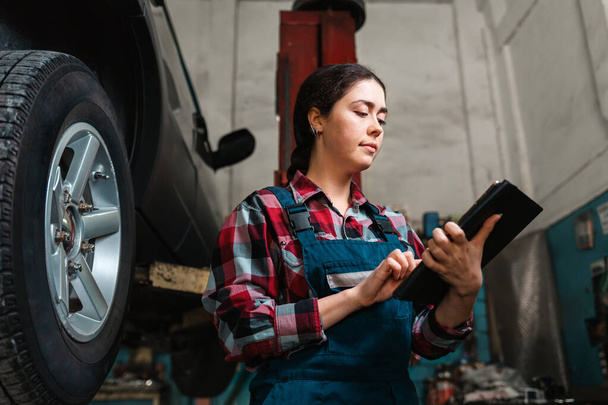 Porträt einer jungen kaukasischen Mechanikerin in Uniform mit einem Tablet in der Hand. Das Rad des Autos auf der rechten Seite. Ansicht von unten. - Foto, Bild