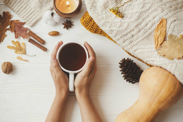 Hands holding warm cup of tea on background of autumn leaves, pumpkin, cozy sweaters, burning candle on white wood. Hello autumn and Happy Thanksgiving. Autumn flat lay - Photo, Image