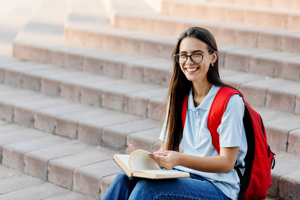 Uma menina bonita estudante de óculos, com uma mochila está sentada nas escadas perto da universidade, lendo um livro, se preparando para palestras ou exames. Formação e educação - Foto, Imagem