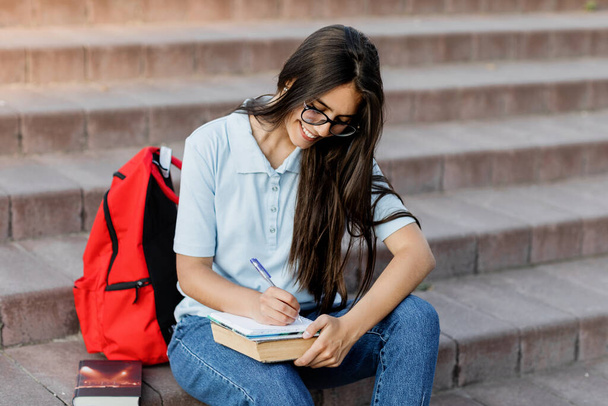 Μια συγκεντρωμένη μαθήτρια με γυαλιά, κάθεται στις σκάλες, γράφει στο σημειωματάριό της. Προετοιμασία για διάλεξη ή εξέταση - Φωτογραφία, εικόνα