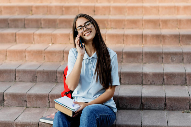 Una ragazza allegra sta parlando su un telefono cellulare con la sua amica, seduta con un taccuino sulle scale del campus - Foto, immagini