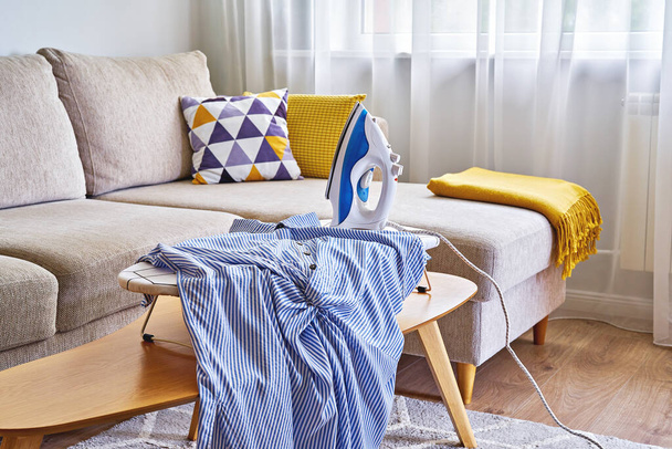 Washed blue shirt on tabletop ironing board and iron in small living room with sofa - Photo, Image