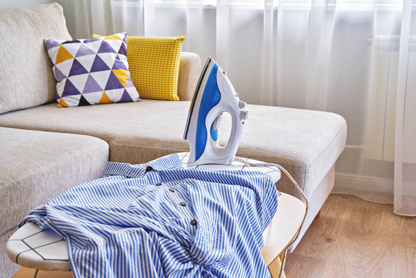 Washed blue shirt on tabletop ironing board and iron in small living room with sofa - Photo, Image