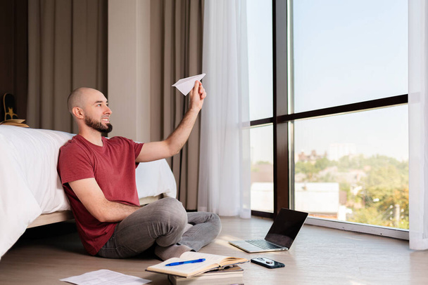 A happy caucasian man sits on the floor and launches a paper airplane into the air. On the floor is a laptop, books and papers. The concept of online courses and online messengers - Photo, Image