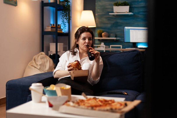 Женщина отдыхает на диване, держа вкусный бургер, пока пьет пиво, смотря развлекательный фильм - Фото, изображение