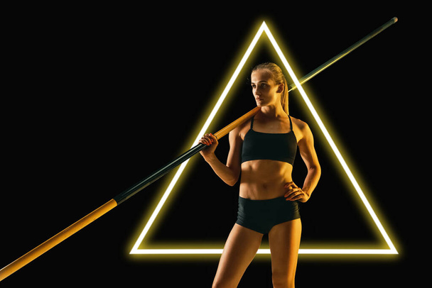 Young female pole vaulter standing isolated on black background with luminescent triangle. Abstract desing, concept of active lifestyle, beauty, motion in sport - Foto, immagini