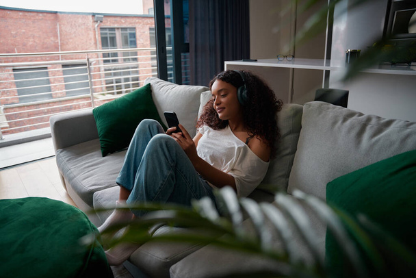 Mixed race female teen with afro relaxing on couch with cellular device  - Φωτογραφία, εικόνα