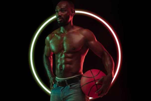 Abstract desing. Young african man, muscular athletewith basketball ball isolated on black background with neon luminescent geometric circle - Fotoğraf, Görsel