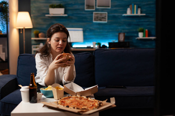 Женщина сидит на диване и ест вкусный бургер, смотрит документальный фильм по телевизору. - Фото, изображение