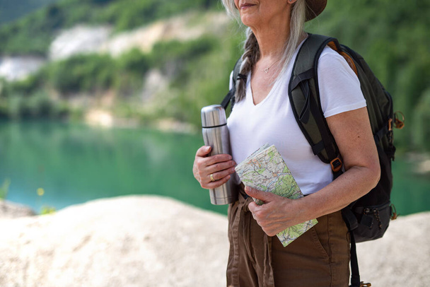 Tanımlanamayan yaşlı kadın turist yaz tatilinde yürüyüş yapıyor, gölün kenarında duruyor.. - Fotoğraf, Görsel