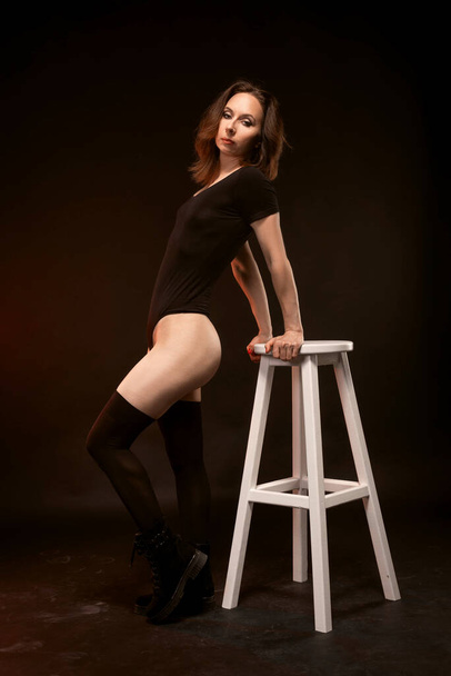 A slender woman in a black bodysuit and stockings poses on a black background near a white stool. Sexy young woman. - Foto, afbeelding