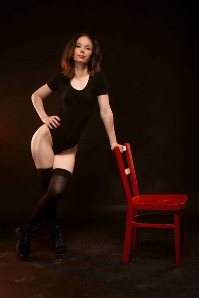 A slender woman in a black bodysuit and stockings poses on a black background near a red chair. Sexy young woman. - Zdjęcie, obraz