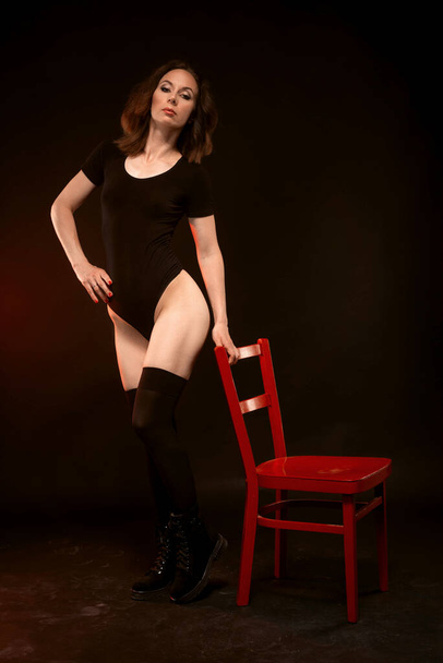 A slender woman in a black bodysuit and stockings poses on a black background near a red chair. Sexy young woman. - Foto, afbeelding