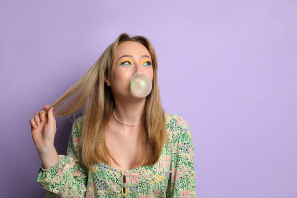 Fashionable young woman with bright makeup blowing bubblegum on lilac background, space for text - Photo, image