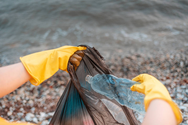 Az önkéntes kezének közelsége gumikesztyűben egy koszos műanyag palackot rak a szemetesbe. Óceán vize a háttérben. Első személyű nézőpont. A környezetszennyezés fogalma. - Fotó, kép