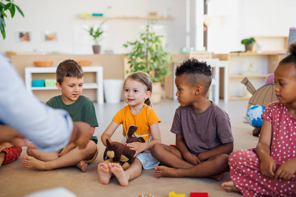 Group of small nursery school children sitting on floor indoors in classroom, listening to teacher. - Фото, изображение