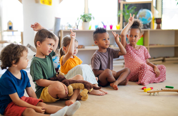 Group of small nursery school children sitting on floor indoors in classroom, raising hands. - Фото, зображення