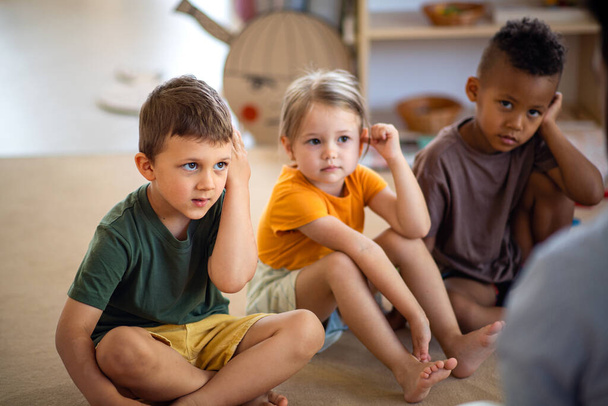 Group of small nursery school children sitting on floor indoors in classroom, listening to teacher. - 写真・画像
