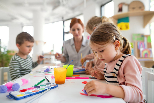 Group of small nursery school children with teacher indoors in classroom, painting. - 写真・画像