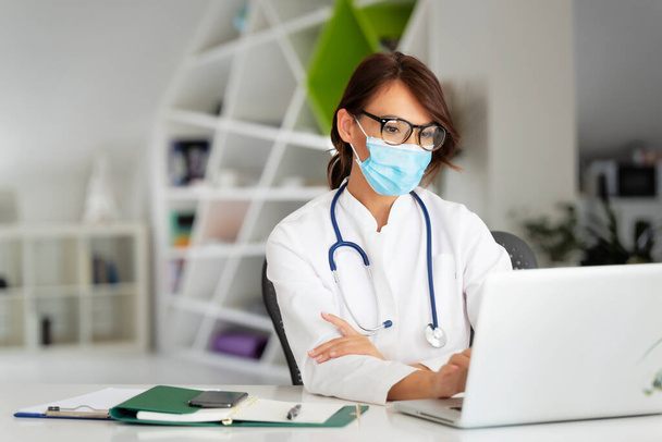 Female doctor wearing face mask while sitting at desk and using laptop - 写真・画像
