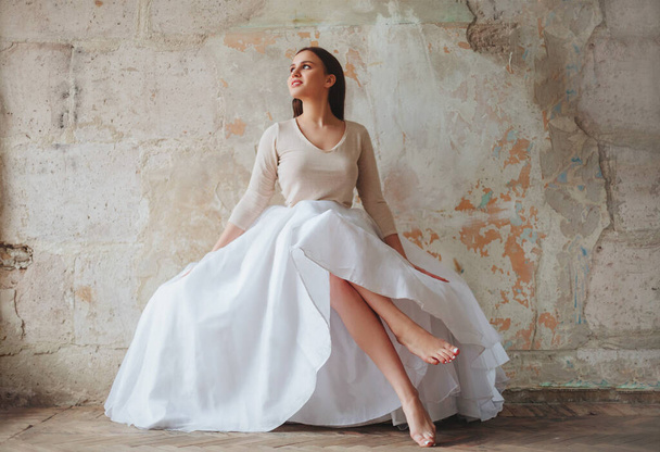 Beautiful young brunette romantic woman sitting barefoot in fluffy long white skirt against concrete vintage style wall in loft beige room, looking aside. Full-length shot - Fotó, kép