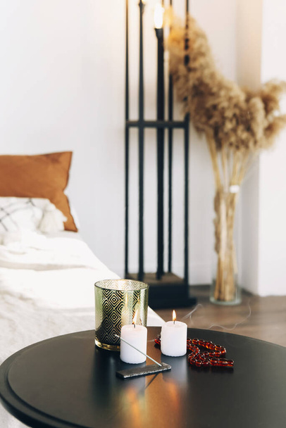 Cropped photo of cozy bedroom interior. Round table with burning candles, slowly smoldering incense on wooden stand and rosary beads. Relax and meditation mood at home for better sleep - Photo, Image